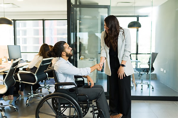 A woman in a wheelchair shaking hands with a man in an office, demonstrating inclusivity and professionalism.