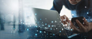 A marketing professional is multitasking, utilizing both a laptop and a cell phone to enhance the reputation of a collection agency.