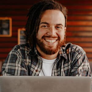 A man with dreadlocks smiles while using a laptop.