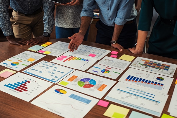 People standing around a table with various charts, graphs, and colorful sticky notes.