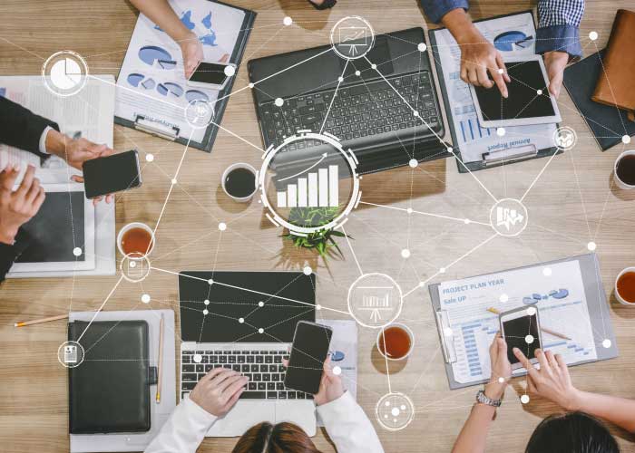 Team collaborating at a desk with laptops, smartphones, charts, and graphs, overlaid with a digital network graphic.