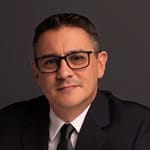 A man in a suit and glasses against a dark background.